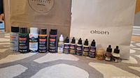 12 bottles of assorted craft and model paints arranged on carpet with two brand bags in background