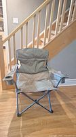Green foldable camping chair open, showing fabric construction, zipper pockets in arm rests, and cup holder on one side.