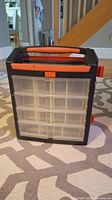 Front view of the screw organizer showing the multiple small clear plastic compartments behind a black and orange frame with an orange latch and carrying handle at the top.