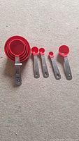 Photo of a set of four red measuring cups nested together and four red measuring spoons with silver handles laid out on a beige carpet surface.