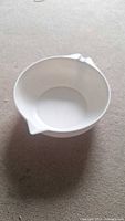 White mixing bowl with handle and spout photographed from above and side angles on carpeted floor.