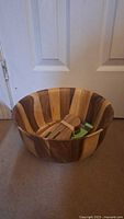 Large wooden salad bowl with visible alternating light and dark wood stripes, placed on carpet in front of white door.