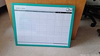 Whiteboard calendar with a 1 month grid layout, turquoise frame, placed leaning against wooden furniture on carpet. Two round magnets visible in top right corner.