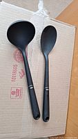 Two black plastic kitchen utensils displayed side by side on cardboard, ladle and serving spoon with hanging holes and silver ring details on handles.