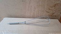 Metal kitchen whisk shown against a cardboard background, laid horizontally, highlighting the bent wire loops on the right end.