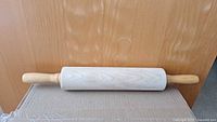 Wooden rolling pin photographed on a cardboard surface against a wood background, showing the full length and smooth cylindrical shape with handles.
