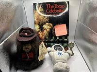 Full lot photo showing brown plush beaver toy wearing a brown hat, white plush robot toy, Expo Celebration book, and dinner bell medal.
