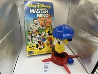 Photo showing Walt Disney Master Mind board game box with colorful Disney character artwork, Mickey Mouse figurine in yellow raincoat holding blue umbrella standing on red base in front of the box.