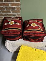 Front view of pair of square Turkish wool kilim-style ottomans with red, black, and multicolor woven patterns, braided edges, placed against brick wall.