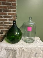 Two large vintage glass wine jugs, one green and one clear, side by side on a white cloth with a brick wall background.