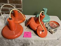 Four mid-century modern style pendant lamps, three retro orange and one turquoise, laid on fabric surface showing cords and mounting parts for sale.