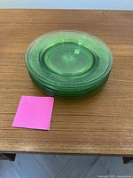 Stack of seven greenish translucent uranium glass side plates on wooden table with pink square for scale.