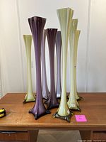 Eight tall Italian glass sculptures arranged on wooden table, showing yellowish-green and purple colors with flared bases.