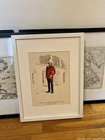 Close-up frontal image showing the white-framed infantry print featuring a soldier in red uniform standing in front of a building. Frame lacks glass and print is matted.