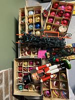 Overview of multiple boxes filled with approx. 70 vintage glass Christmas ornaments in various colors, 8 light reflectors, Christmas light strings, and wooden nutcracker figure.