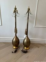 Pair of vintage-style table lamps with bulbous gold-toned bases, tall necks with tassel cords, and dual light sockets with clear textured bulbs, shown against a white wall and wooden floor.