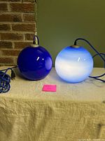 Two blue spherical glass pendant lights sitting on a white cloth, one illuminated showing a white glow with blue edges, the other unlit.