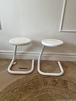 Front view of the pair of white tubular metal Haworth Paperclip stools showing their cantilevered paperclip shape and round seats on a wooden floor with woven mat.