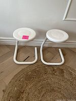 Two white metal Haworth Paperclip stools seen from front, showing round seats and bent tubular bases with minor surface wear.