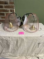 Two pink glass pendant lamps with scalloped ridge design, gold bases, and black cords displayed side by side on cloth-covered surface.