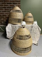 Set of 3 natural rattan woven pendant light shades displayed on a white cloth against a brick wall and green backdrop with two white cord kits beside.