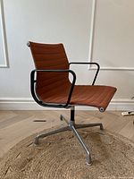 Side profile of rust orange Eames Aluminum Group chair on round jute rug, showing aluminum base and frame