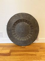 Front view of vintage metal round decorative plate showing scalloped floral embossed pattern and dark aged patina, placed against wall on hardwood floor.