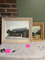 Two framed amateur paintings side by side on a table, showing their appearance and size relative to a marker and tape measure.