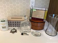 Image showing all eight household items on a countertop against a hex tile backsplash: glass decanter, stained glass frame, tissue box, wooden box, glass vase, metal turtle, ashtray, tea tin.