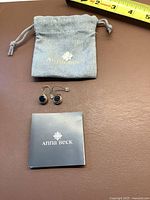 Anna Beck branded grey drawstring pouch with pair of silver earrings and small booklet on brown surface with measuring tape visible.