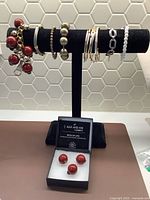 Seven diverse bracelets on black display stand alongside boxed set of four red bead stud earrings.
