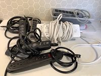 Photo showing 8 surge protectors in varied colors and outlet counts, with cords coiled and some power switches visible.
