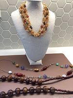 Photo showing three assorted necklaces, including a multi-strand chip bead necklace on a display bust and two other bead necklaces laid flat.