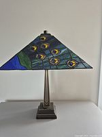 Front view unlit lamp showing square peacock feather stained glass shade and bronze-tone metal base