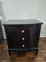 Front view of one dark brown 3-drawer nightstand showing metal handles and curved drawer fronts on wood parquet flooring.