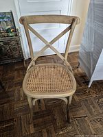 Front view of the vintage wood chair showing cross-back design and cane seat, placed on parquet flooring near a stained glass cabinet and shrink-wrapped item.