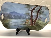 Porcelain serving tray showing hand painted nature scene with two deer near trees, mountains and water in background, set on a display stand to highlight design and shape with handle cutouts