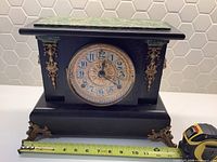 Front view of the vintage Seth Thomas mantel clock showing the faux marble top, brass decorations, clock face and part of a tape measure for scale.