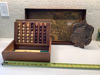 Wooden box containing a vintage board game with light and dark wooden balls arranged in rows, placed next to a wooden decorative tray featuring a painted house and trees scene, and a small plaque.