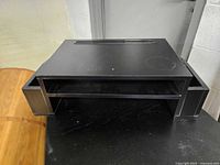 Front view of black computer work station showing top surface with slot and side drawer compartments pulled out slightly.