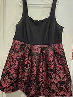 Front view of sleeveless dress with solid black top and dark red and black damask patterned skirt on hanger against white wall.