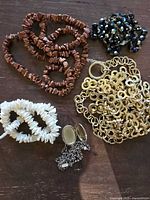 Photo showing five items: brown irregular bead necklace, black faceted bead necklace, white shell bead necklace, gold-tone interlocking link chain belt, and silver-tone locket pendant on chain.