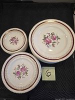 Three sizes of floral porcelain plates stacked, showing decorative rose pattern and burgundy-gold border, with sticky note '6' nearby