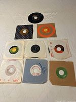 Photo showing nine 45 rpm vinyl records in various sleeves arranged on a quilted surface.