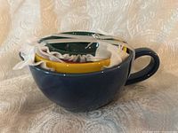 Stacked set of four ceramic measuring cups in original packaging, nested in blue cup with a handle showing, rubber bands and tissue secure them.