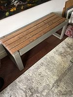 Front angled view of rectangular wooden bench with slatted wood top and grayish solid wood legs against wall with painting and carpet.