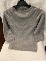 Grey cable knit patterned cashmere sweater hanging on a black hanger, showing overall front view with 3/4 length sleeves and round neckline.