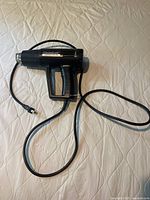Black Hot Shot electronic heat gun with attached power cord laying on white quilted surface, showing overall shape and size.