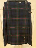 Full view of dark green wool plaid ladies kilt hanging, showing pleating and stitching