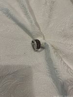 Front and side view of a sterling silver wide band ring with rows of dark gemstones set on the top half of the band, placed on a textured light fabric background.